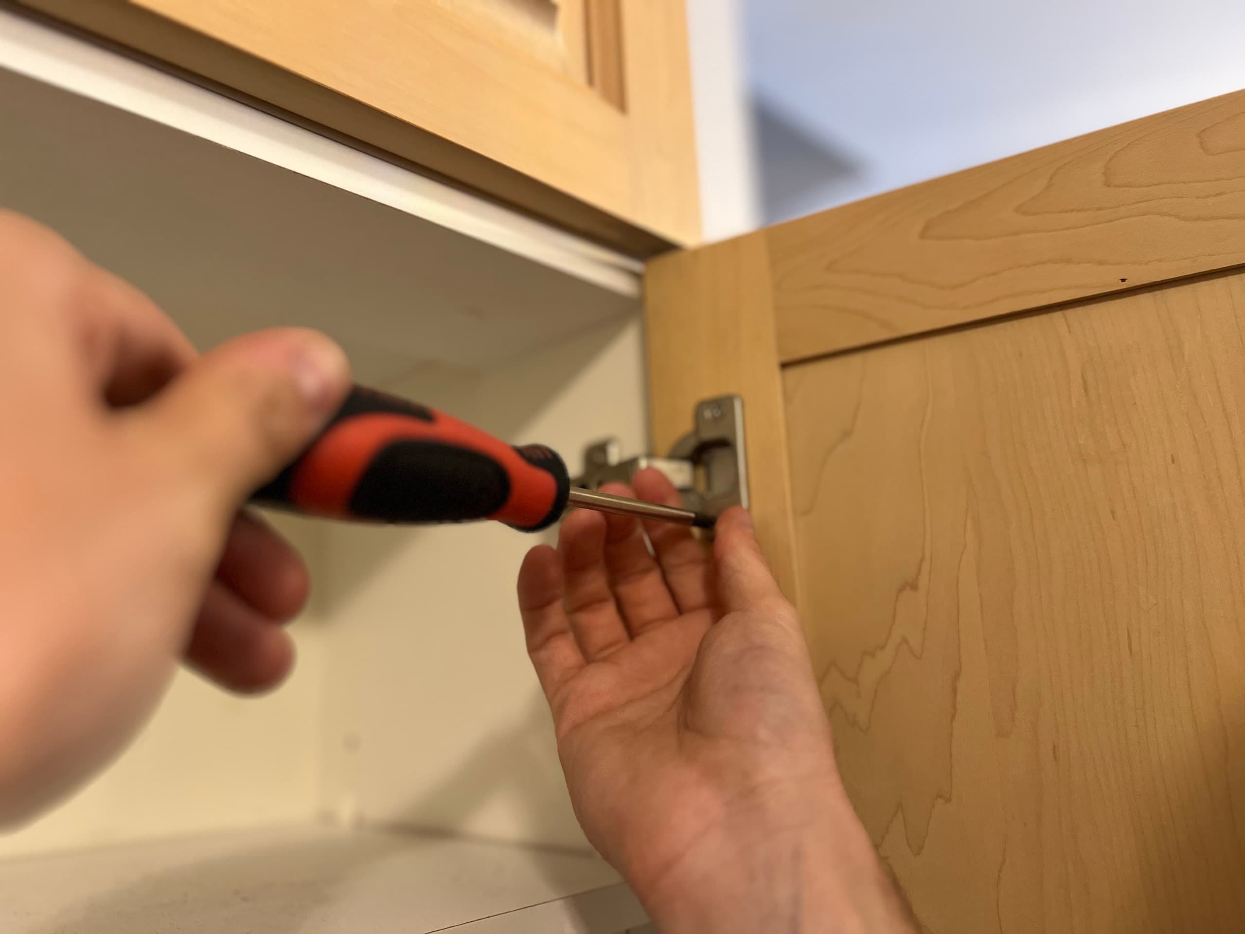 A hinge being unscrewed from a maple cabinet door during disassembly