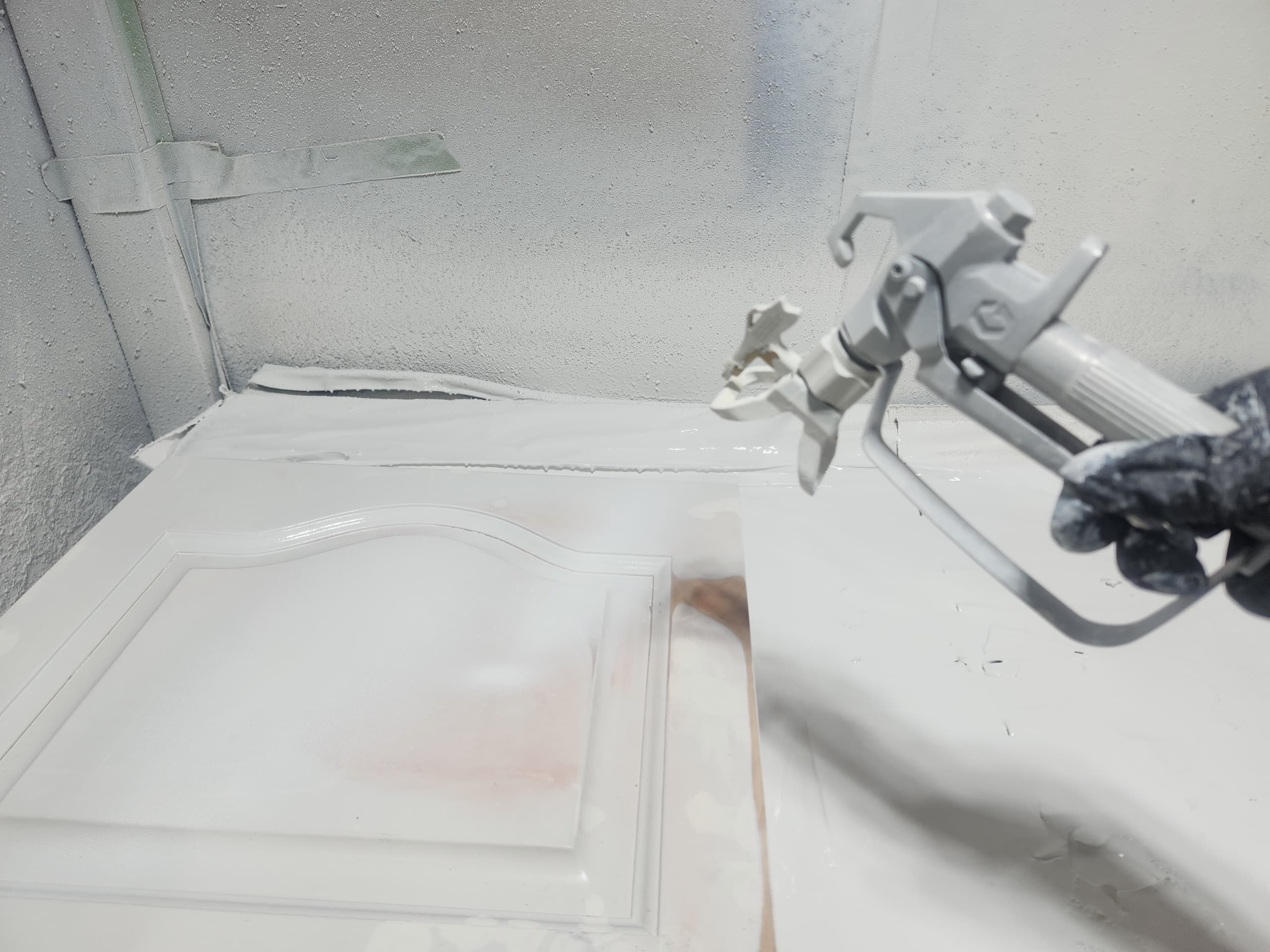 A spray gun laying a wet white coat across a cabinet door lying flat at the shop